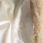 The Nella Letter Necklace with a gold A pendant is displayed on cream satin fabric beside textured dried grass.