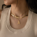 A woman wears layered gold necklaces, including the Nella Letter Necklace with a letter A pendant and a personalized script name necklace.