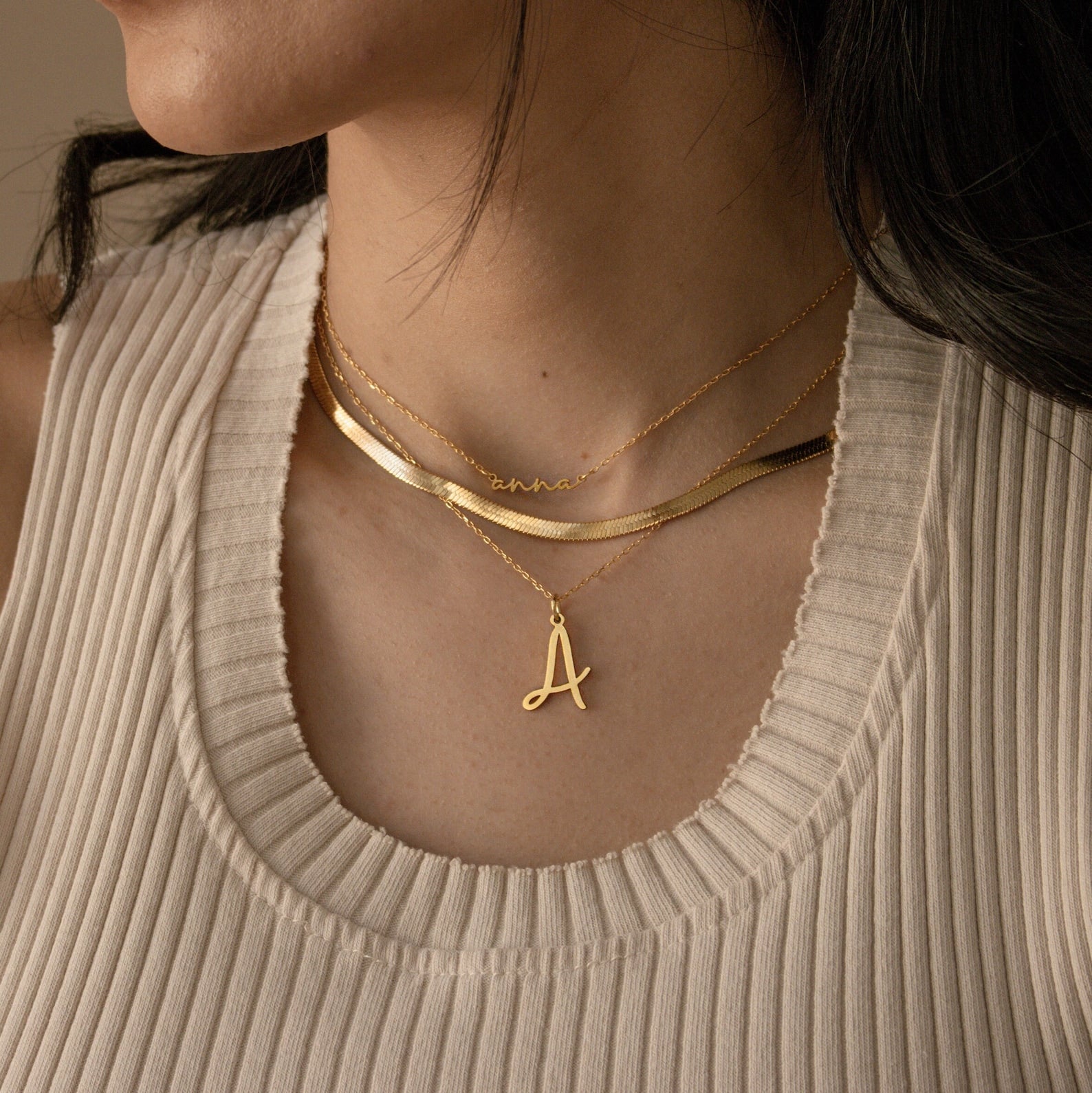 A woman wears layered gold necklaces, including the Nella Letter Necklace with a letter A pendant and a personalized script name necklace.