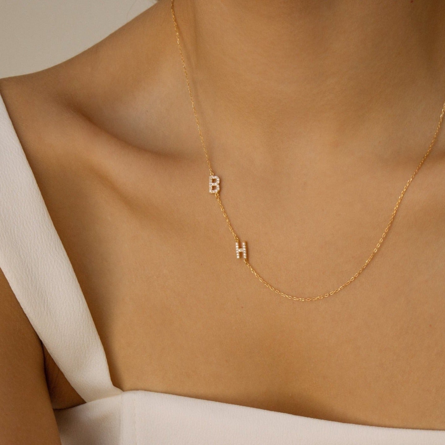 A woman wears the Pave Sideways Initial Necklace with hanging B and H charms over a white off-shoulder top.