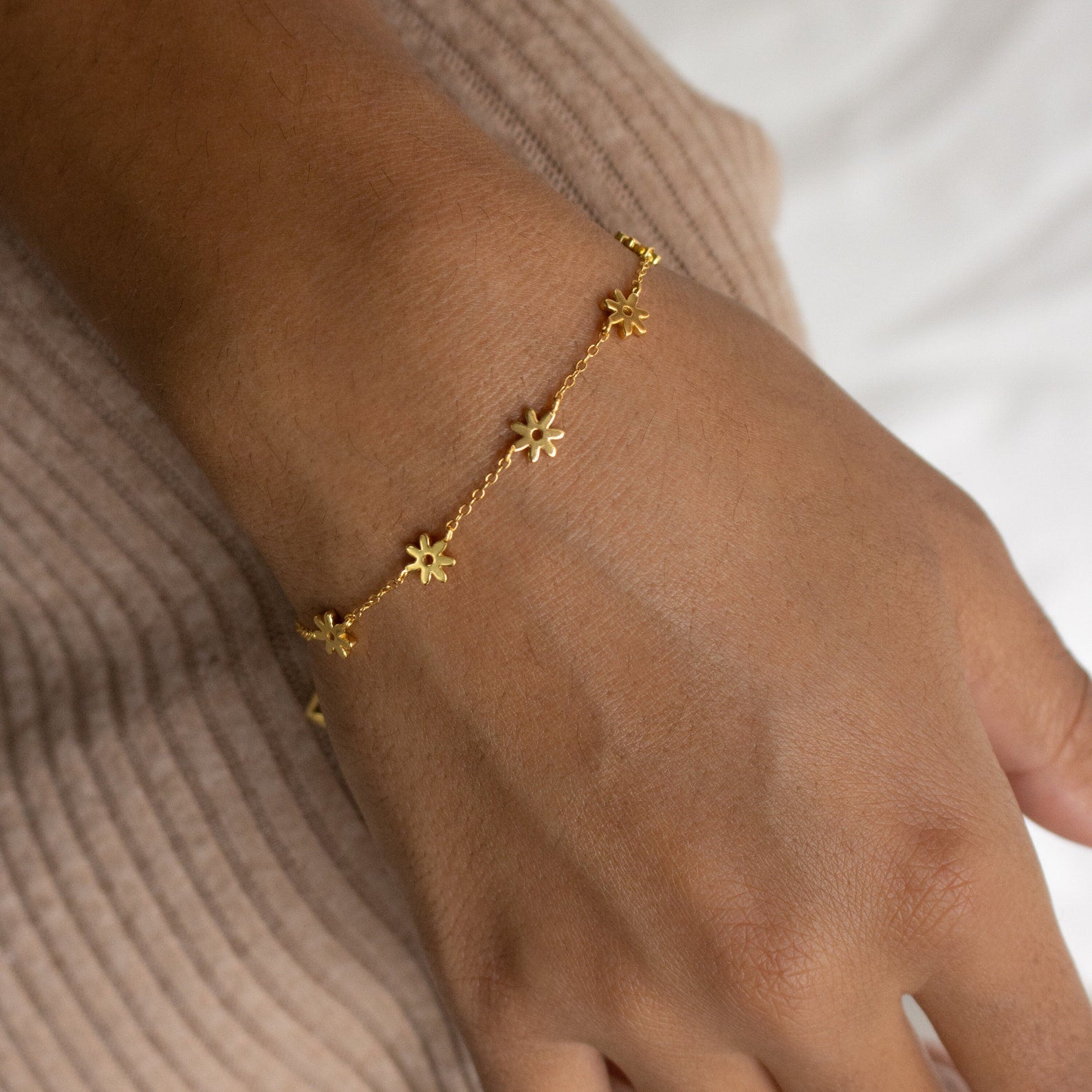 The Flower Child Bracelet showcases gold daisy charms on a wrist, set against beige ribbed fabric—an ideal accessory for cottage-core or boho-hippie style lovers.