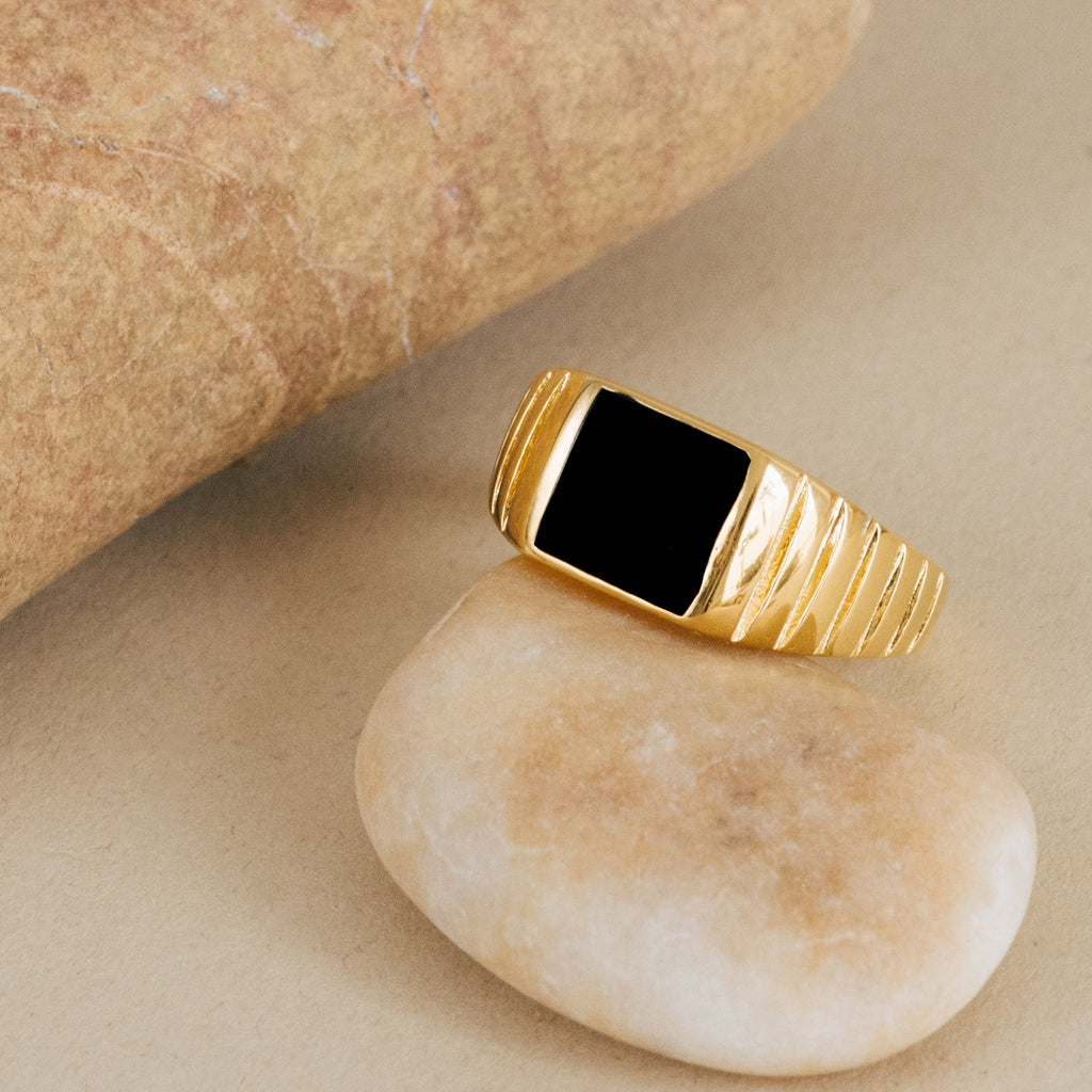 The Black Signet Ring, featuring a black rectangular stone set in gold, is displayed on a smooth beige pebble near a larger rock.