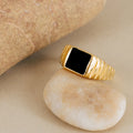 The Black Signet Ring, featuring a black rectangular stone set in gold, is displayed on a smooth beige pebble near a larger rock.