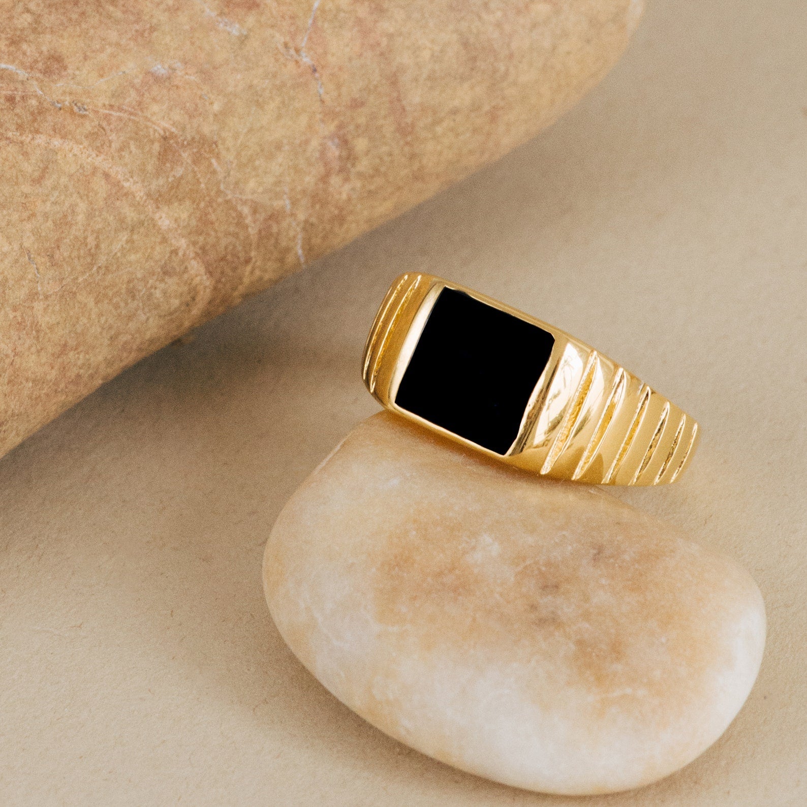 The Black Signet Ring, featuring a black rectangular stone set in gold, is displayed on a smooth beige pebble near a larger rock.