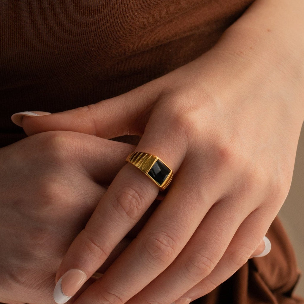 A hand wearing the Black Signet Ring—a unisex gold ring with a striking black rectangular stone—rests gently on another hand.