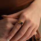 A hand wearing the Black Signet Ring—a unisex gold ring with a striking black rectangular stone—rests gently on another hand.