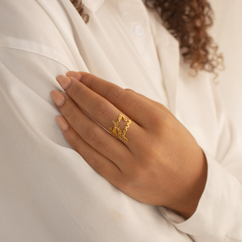 A hand with nude nails showcases the Grace Duo Name Ring in gold, a personalized name ring, resting elegantly on a white shirt sleeve.