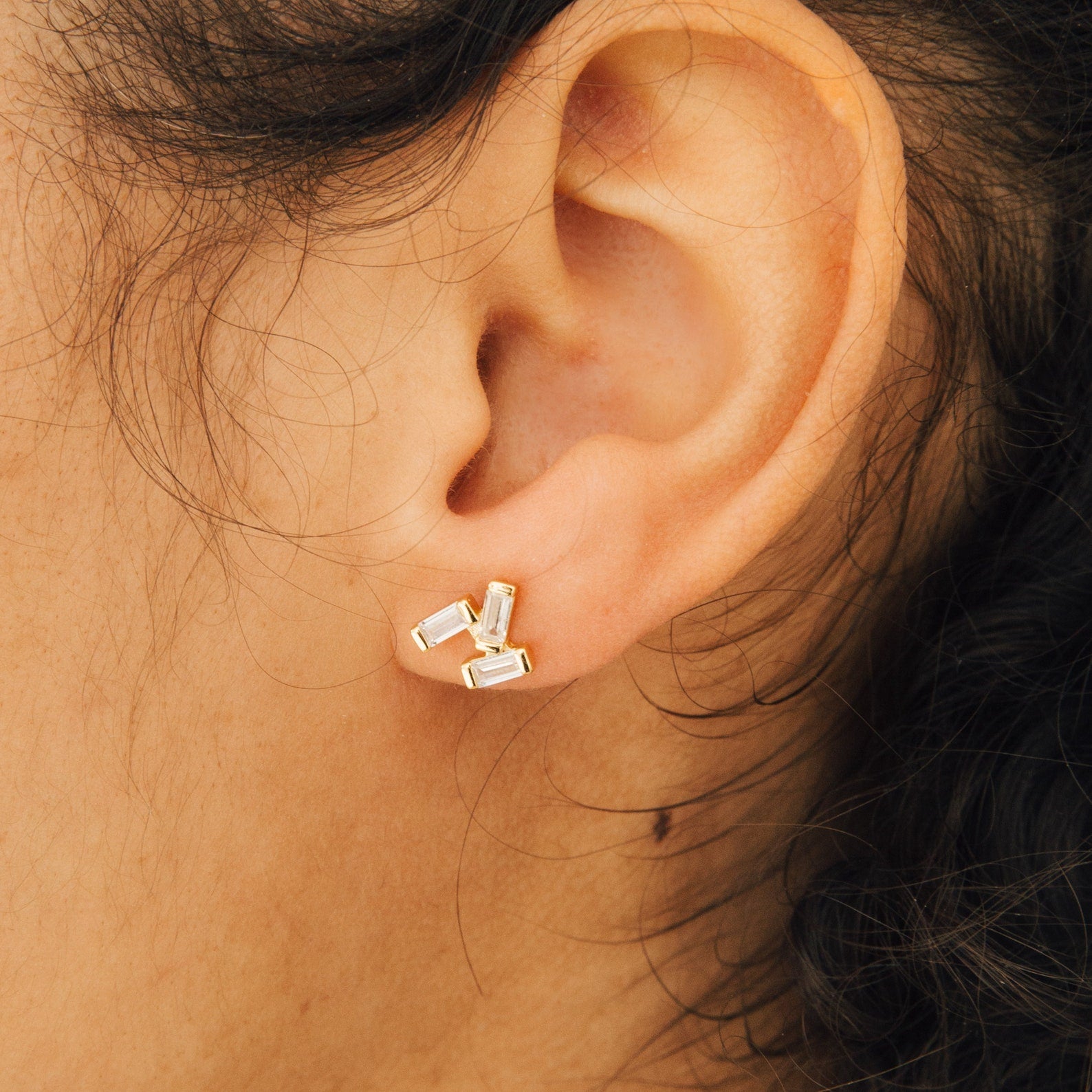 A close-up of an ear wearing the Glacier Diamond Studs, gold earrings featuring three rectangular clear stones.