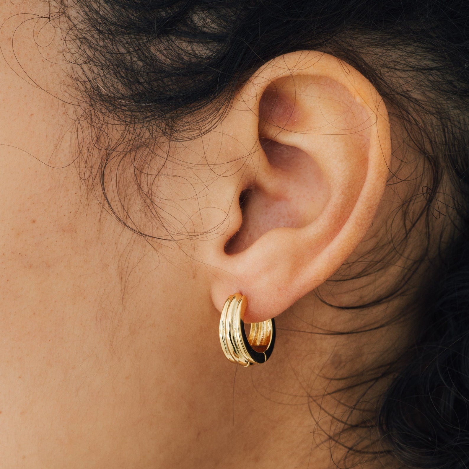 Close-up of a person’s ear wearing Canal Chunky Hoops—small, thick gold earrings with wavy details—perfect contemporary huggies for everyday wear.