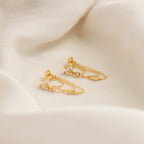 The Genesis Chain Earrings feature gold chains with small crystal accents, making them the perfect statement jewelry piece. Shown on a soft, cream-colored fabric background.