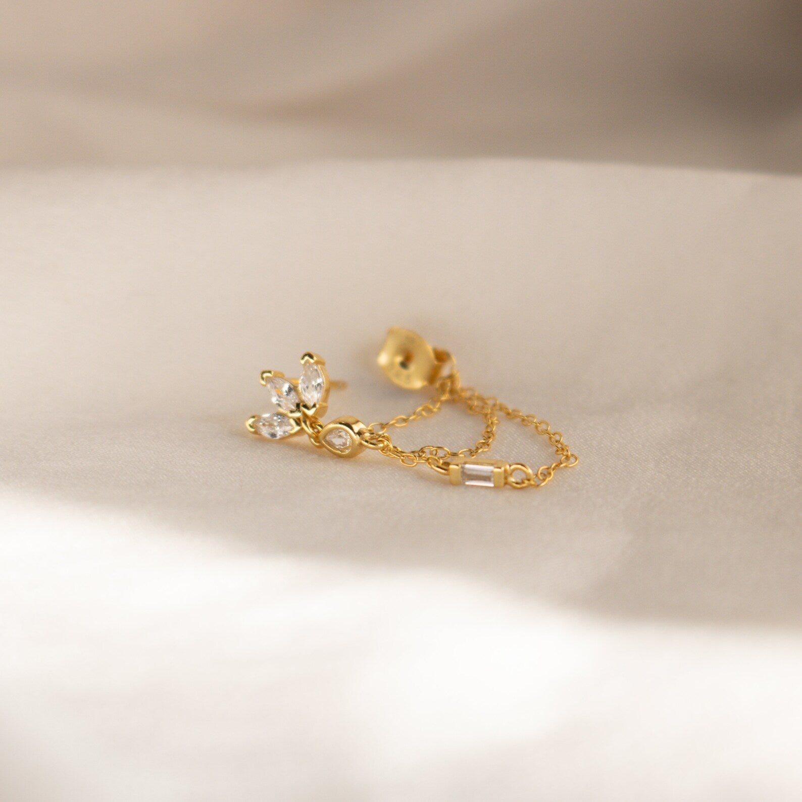 The Genesis Chain Earrings feature a delicate gold chain with three small clear gemstones, displayed on soft, light fabric—ideal for lovers of elegant chain earrings and subtle statement jewelry.