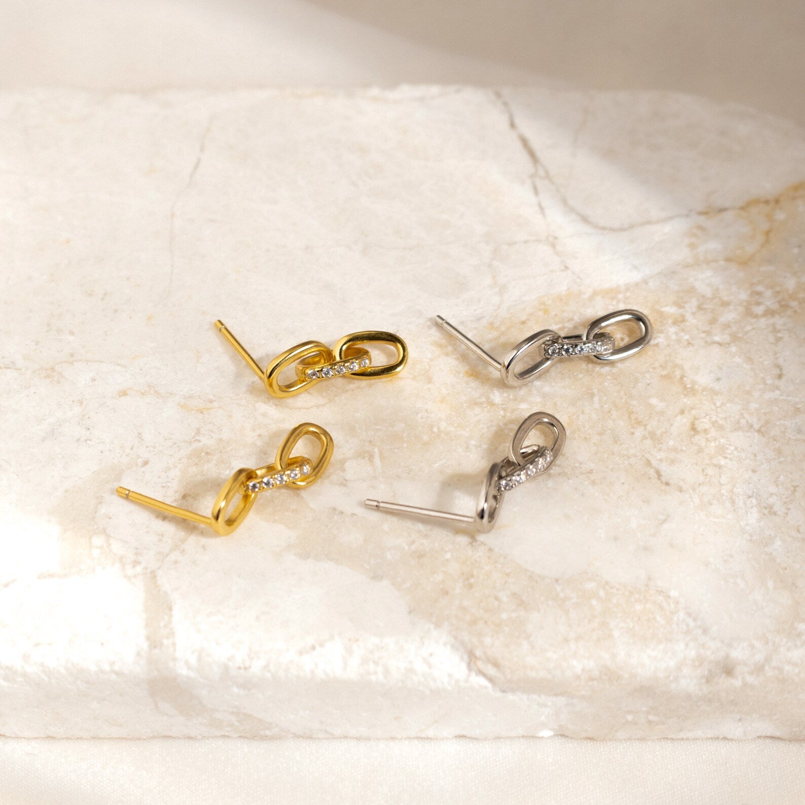 Two pairs of knot-shaped stud earrings—one gold, one silver—rest on textured stone, ideal for pairing with your Amia Pave Link Earrings or adding a subtle pavé touch to any outfit.
