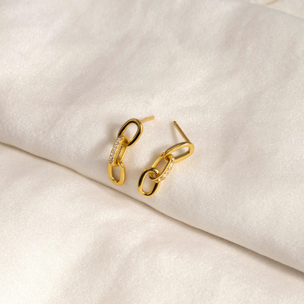 The Linked Earrings Set—gold chain link earrings with small crystals shown on a soft white fabric background—adds a touch of elegance to any modern jewelry collection.