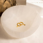 Emily Pave Star Huggies, gold celestial hoop earrings with star charms, rest in a white bowl; a softly blurred book and glass object appear in the background.