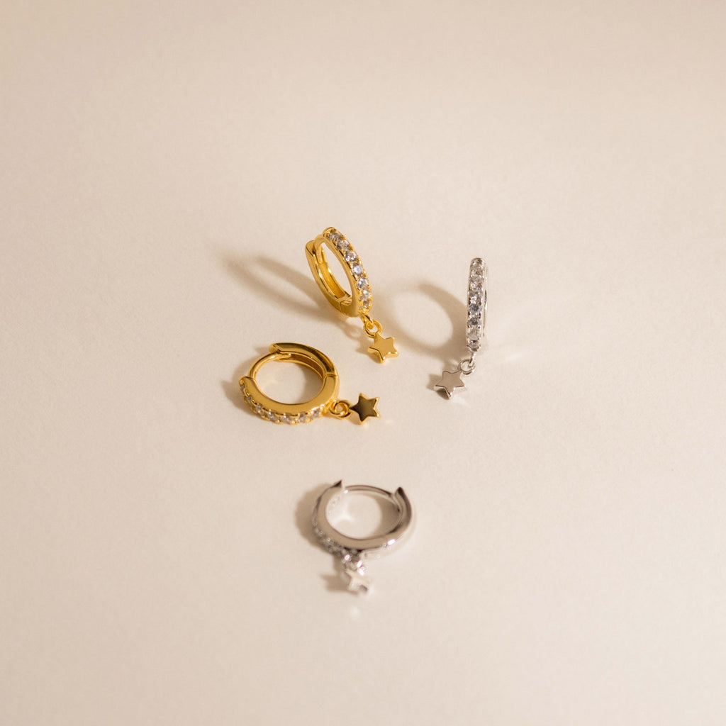 Add celestial sparkle to your style with the Emily Pave Star Huggies—two pairs of hoop earrings in gold and silver, each featuring a dangling star charm. Perfect for shining bright against a beige background.