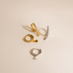 Add celestial sparkle to your style with the Emily Pave Star Huggies—two pairs of hoop earrings in gold and silver, each featuring a dangling star charm. Perfect for shining bright against a beige background.