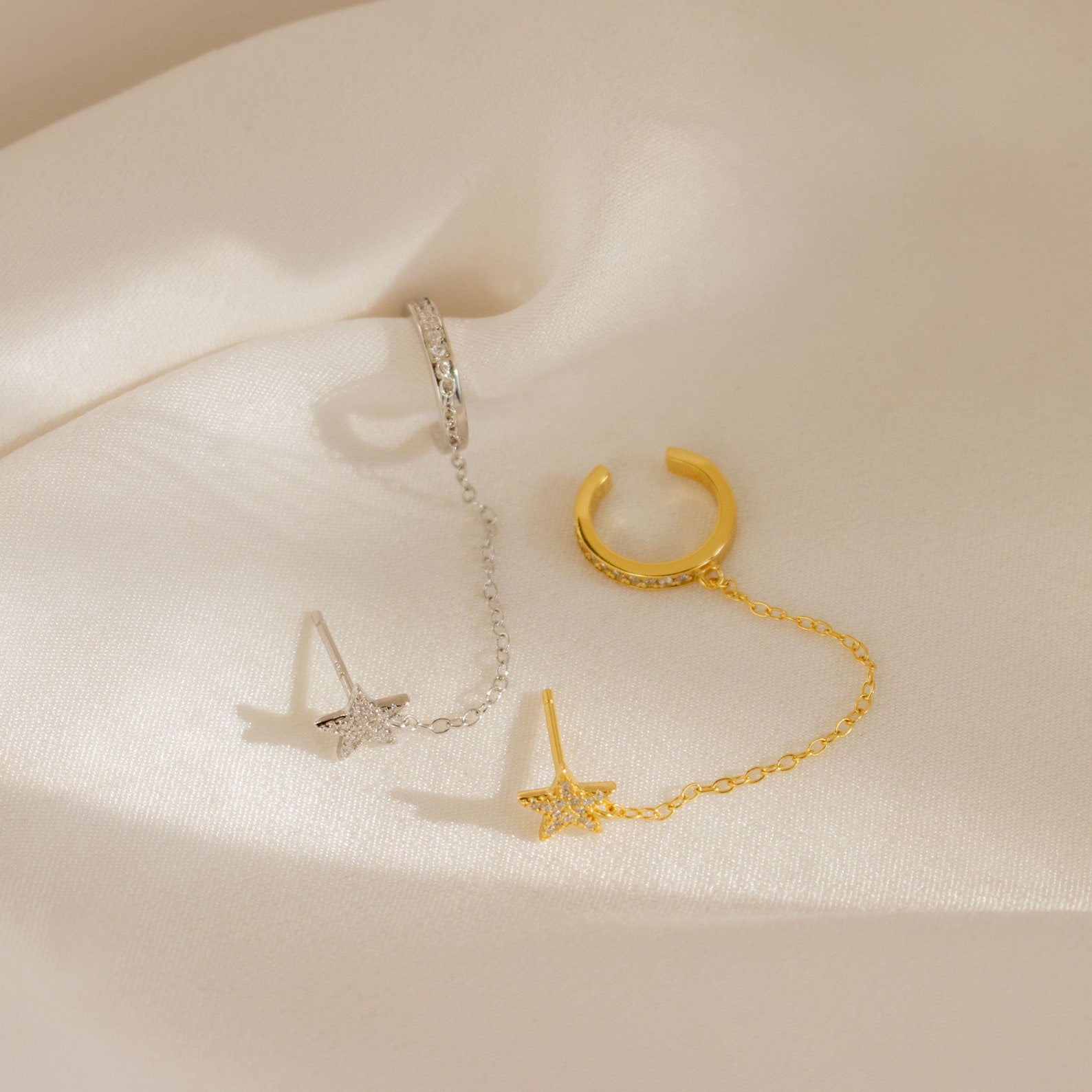 Star Pave Chain Ear Cuffs in gold and silver, featuring diamond-studded star charms and delicate chains, are elegantly displayed on soft cream-colored fabric.
