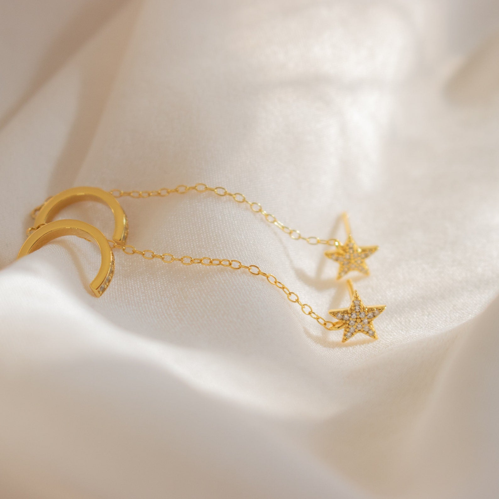 Gold chain earrings with small crescent moons and a diamond-studded star charm, similar to the Star Pave Chain Ear Cuffs, are shown on soft white fabric and feature an adjustable cuff for the perfect fit.