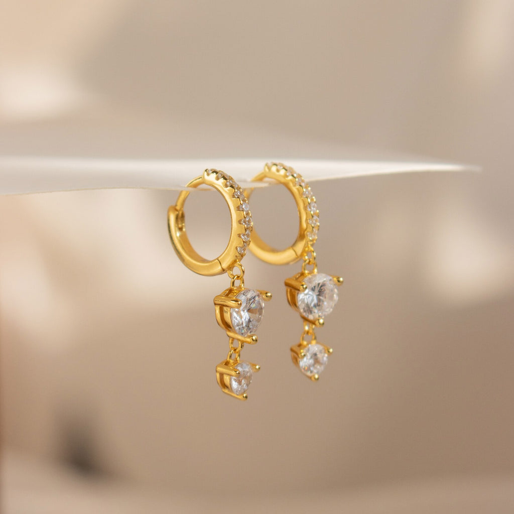 The Caroline Pave Drop Huggies feature gold-embellished hoops accented with small crystals and dangling heart-shaped clear gemstones, shown on a white surface.