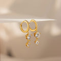 The Caroline Pave Drop Huggies feature gold-embellished hoops accented with small crystals and dangling heart-shaped clear gemstones, shown on a white surface.