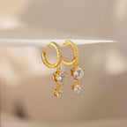 The Caroline Pave Drop Huggies feature gold-embellished hoops accented with small crystals and dangling heart-shaped clear gemstones, shown on a white surface.