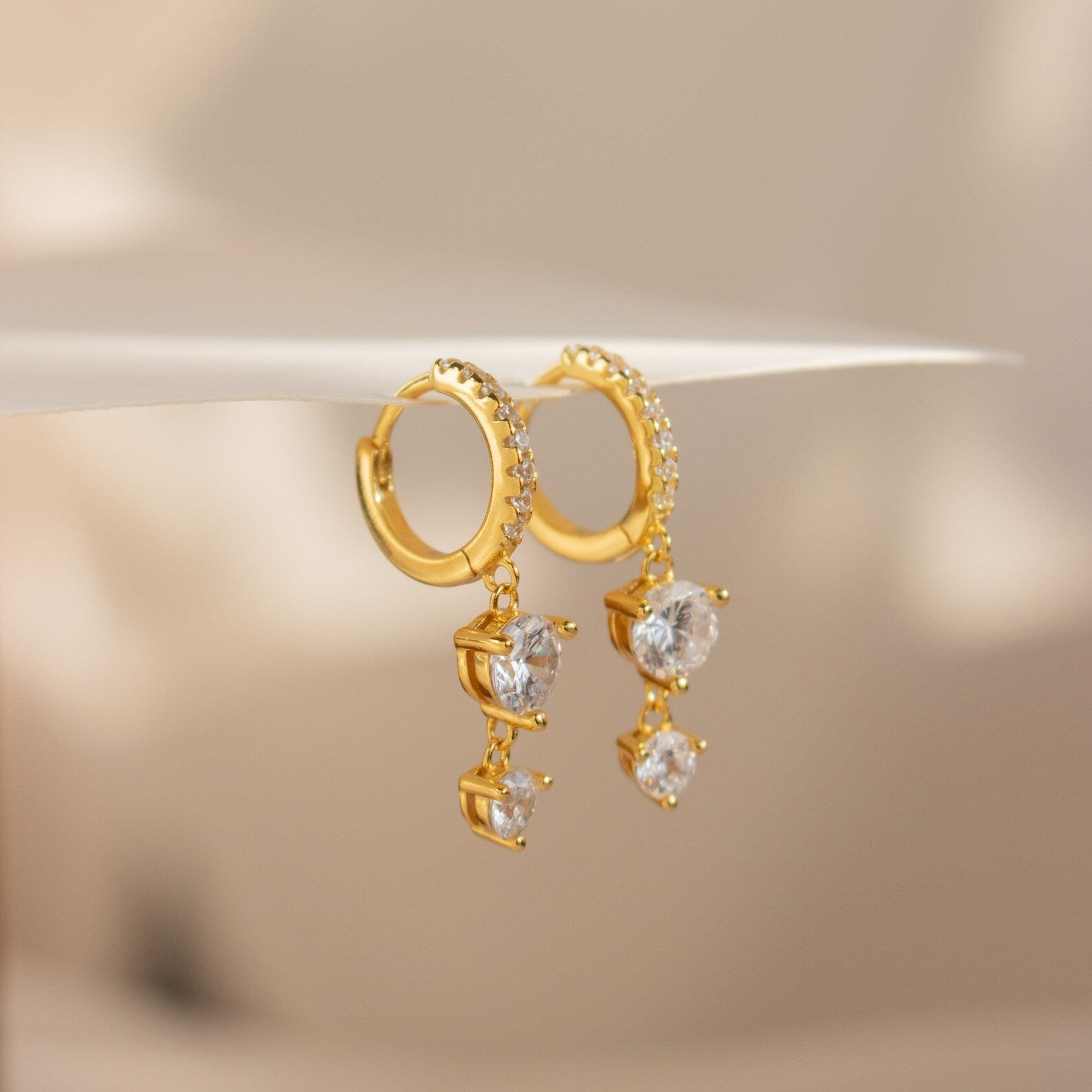 The Caroline Pave Drop Huggies feature gold-embellished hoops accented with small crystals and dangling heart-shaped clear gemstones, shown on a white surface.