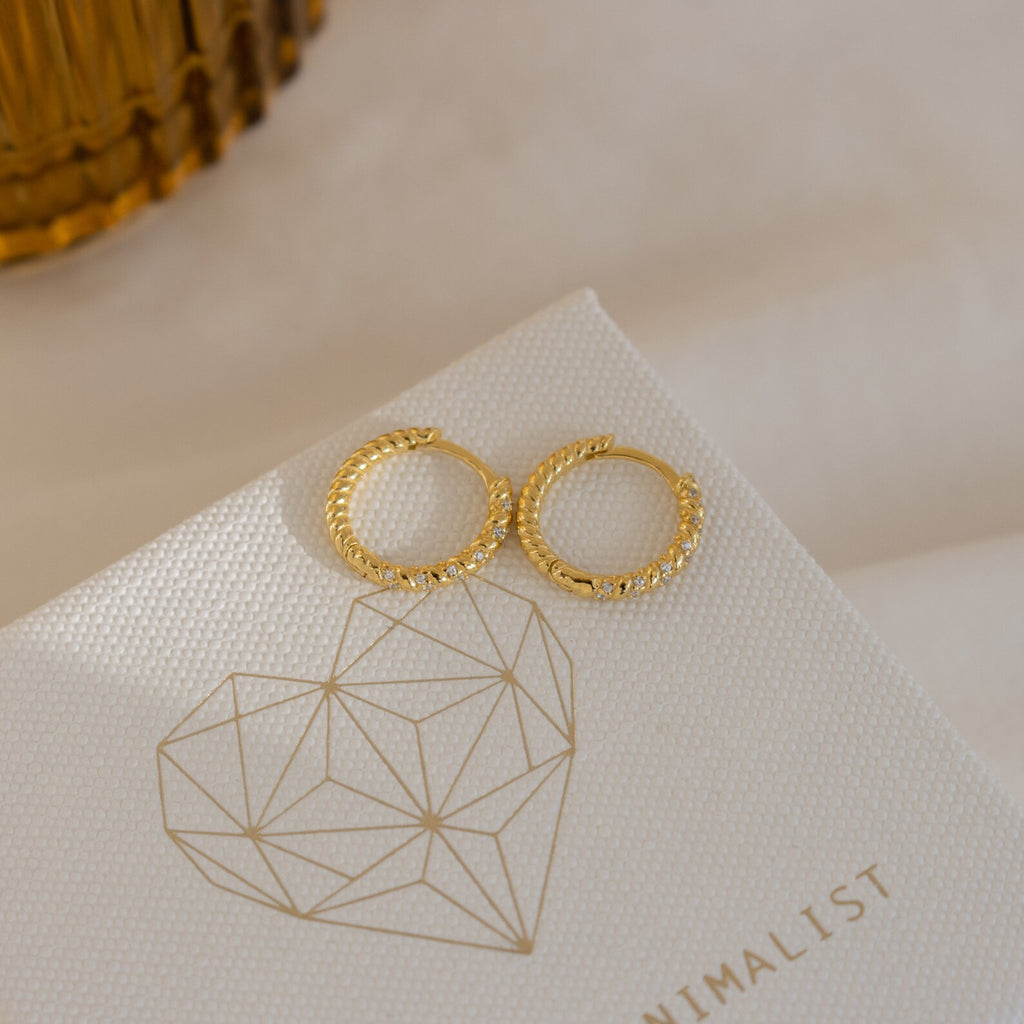 Aster Pave Twisted Hoops—two delicate hoops accented with pavé diamonds—are displayed on a textured card with a geometric heart design.