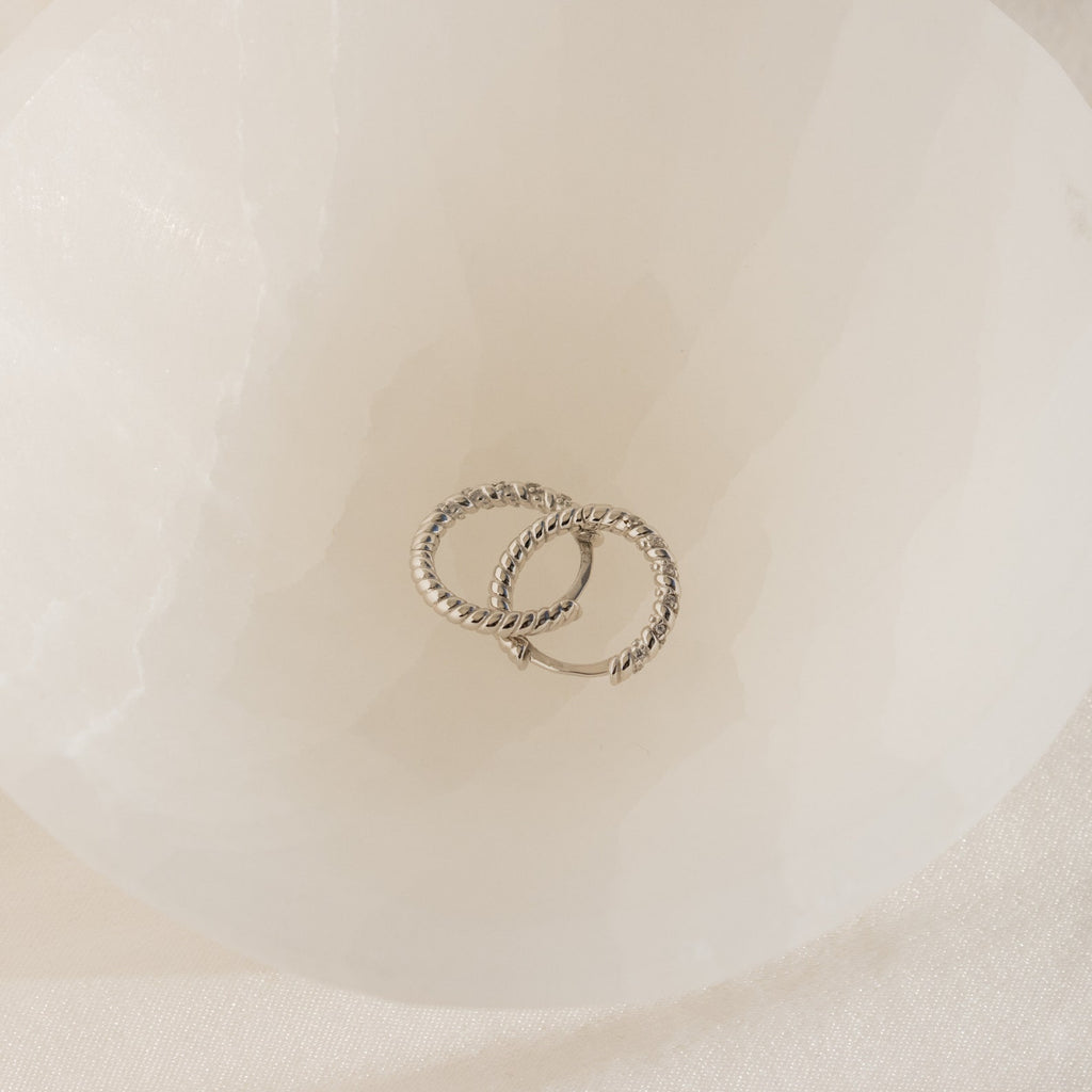 Two Aster Pave Twisted Hoops, crafted in silver and elegantly intertwined, are displayed inside a white, translucent bowl.