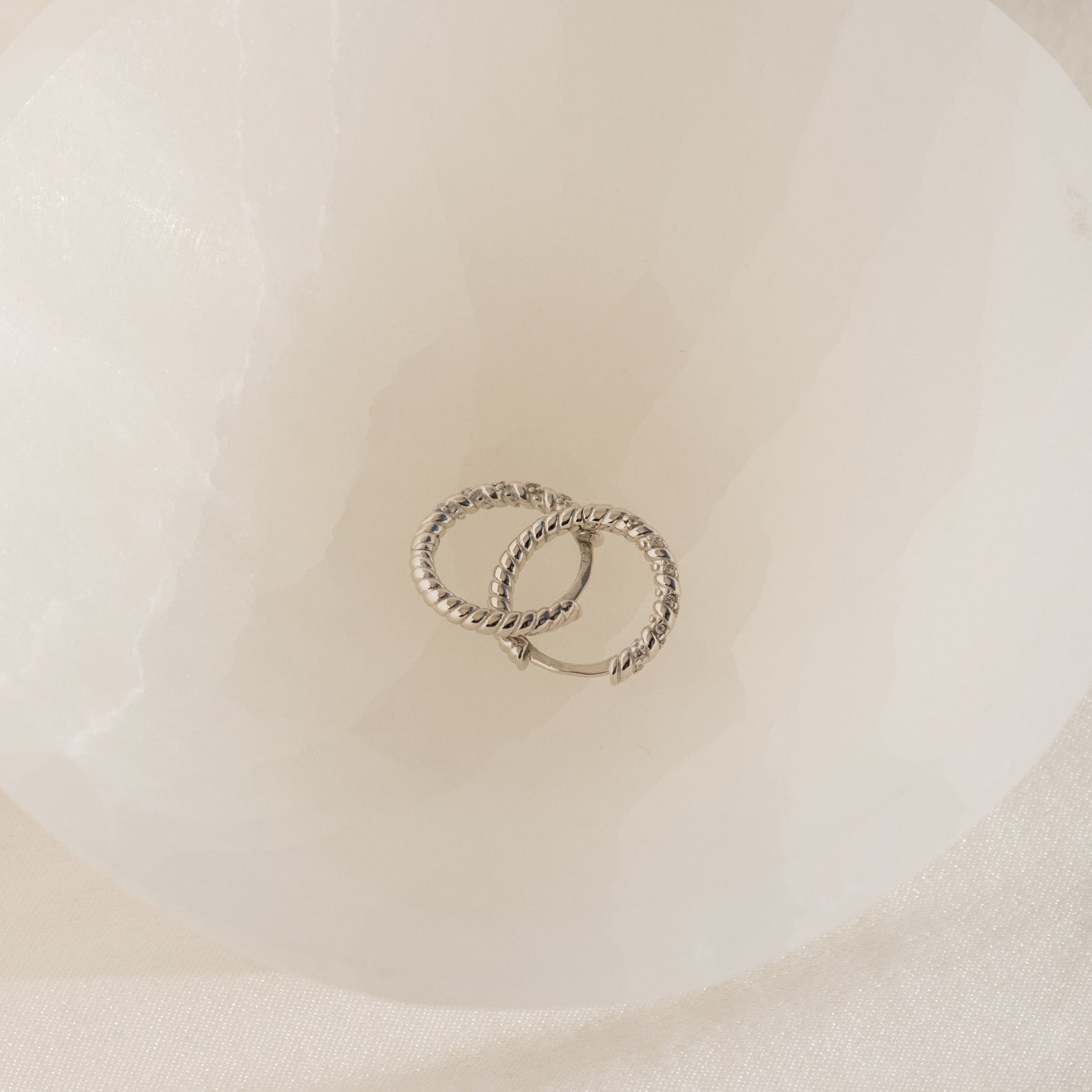 Two Aster Pave Twisted Hoops, crafted in silver and elegantly intertwined, are displayed inside a white, translucent bowl.