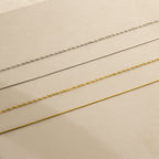 Four delicate necklaces—including the Monet Duo Chain—are arranged in parallel lines on a beige surface.