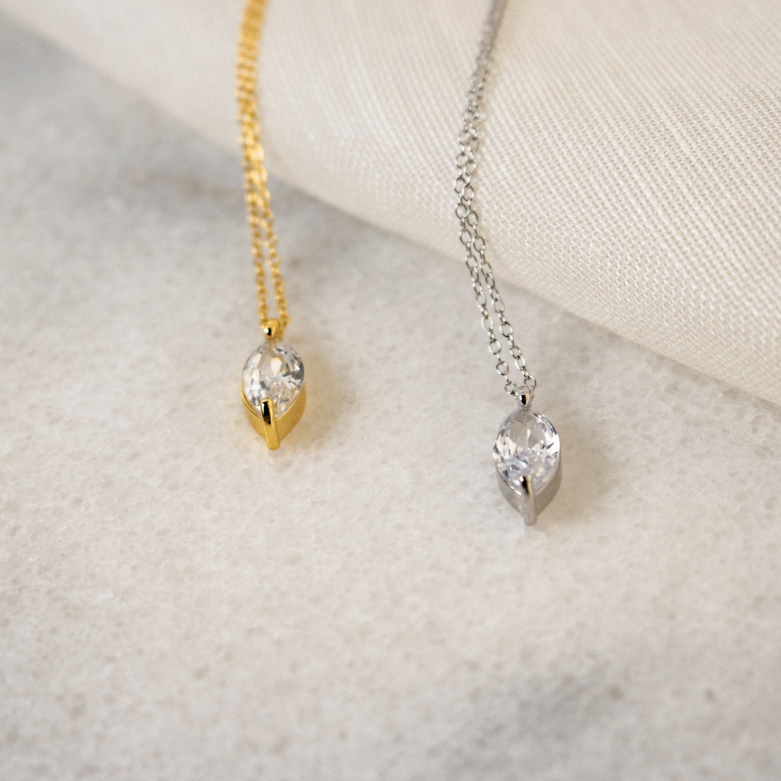 The Athens Diamond Necklace, featuring a marquise-cut crystal pendant on a slender cable chain, is displayed elegantly on light fabric and stone surfaces.