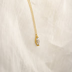 The Athens Diamond Necklace features a gold chain with a clear marquise diamond pendant, shown on a soft cream-colored fabric background.