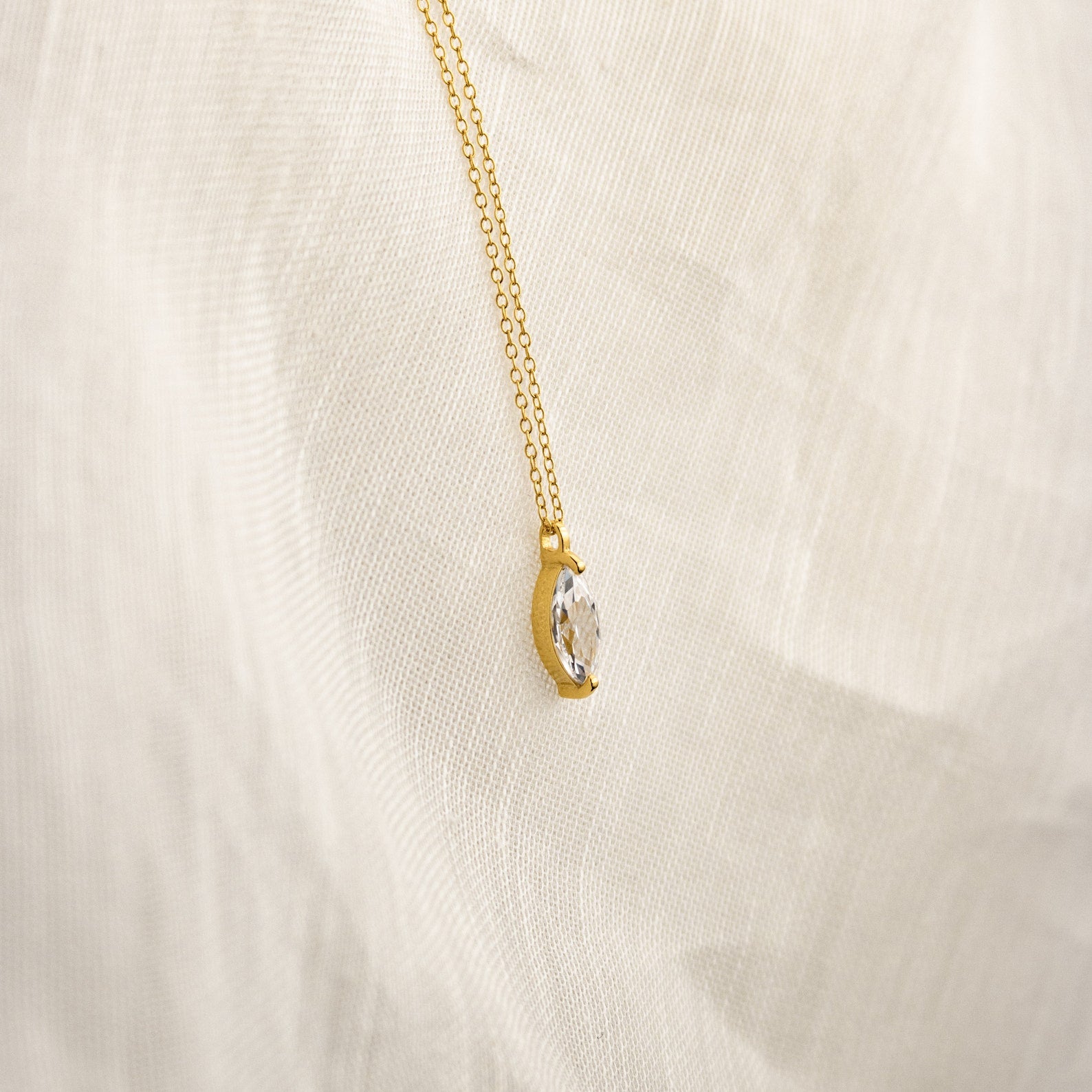 The Athens Diamond Necklace features a gold chain with a clear marquise diamond pendant, shown on a soft cream-colored fabric background.