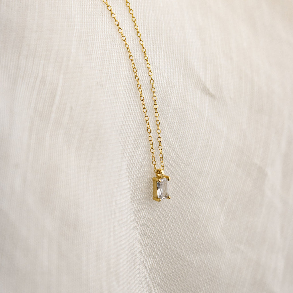 The Sage Diamond Necklace features a gold chain and a rectangular clear gemstone pendant, displayed on white fabric—an effortless touch of timeless elegance.