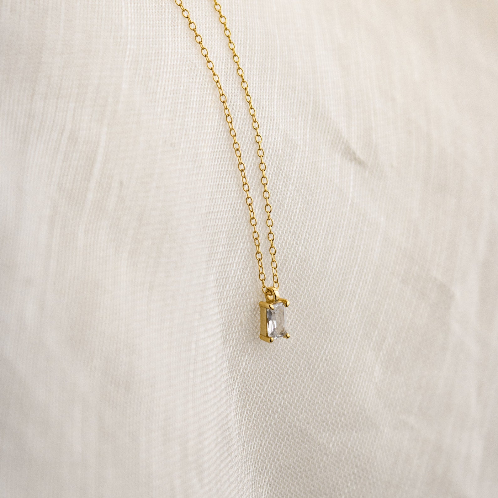 The Sage Diamond Necklace features a gold chain and a rectangular clear gemstone pendant, displayed on white fabric—an effortless touch of timeless elegance.