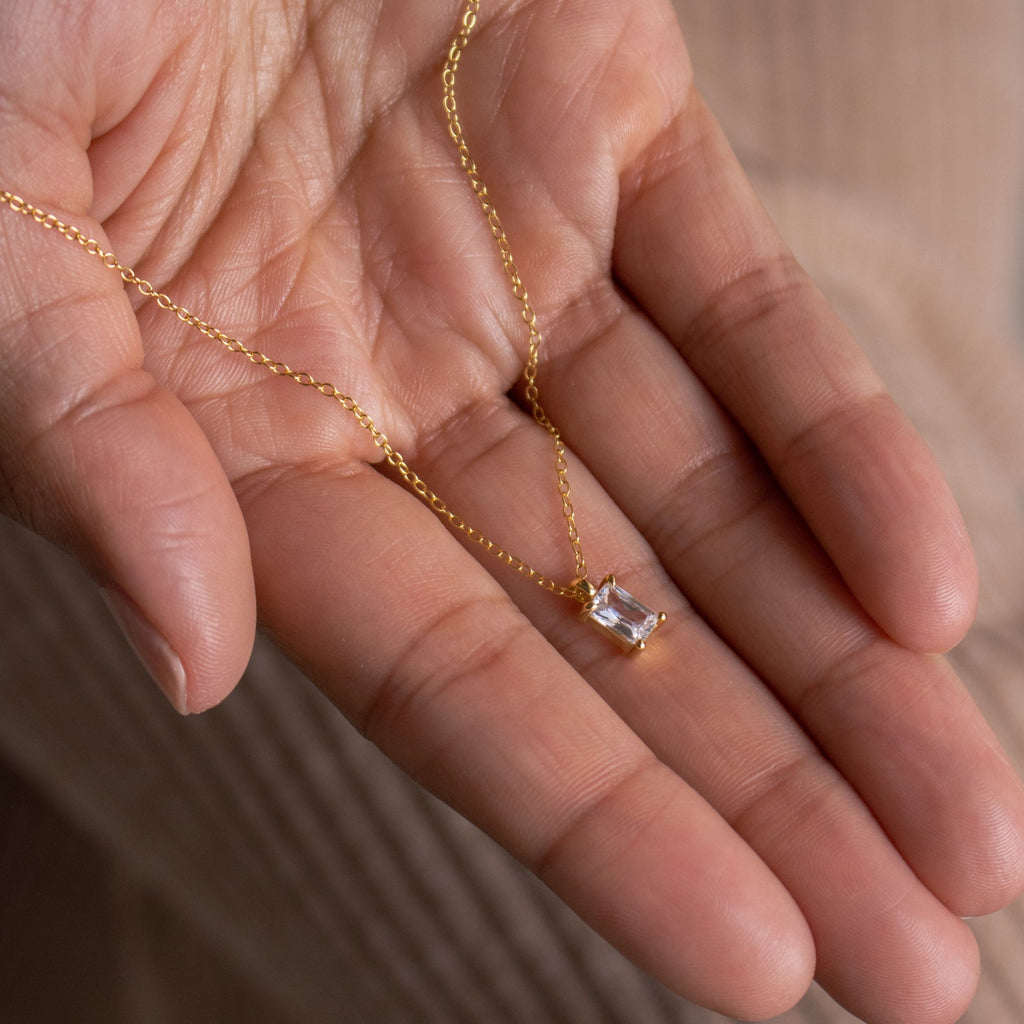 A hand holds the Sage Diamond Necklace, featuring a delicate gold chain with a rectangular diamond pendant, capturing the timeless elegance of luxury jewelry.