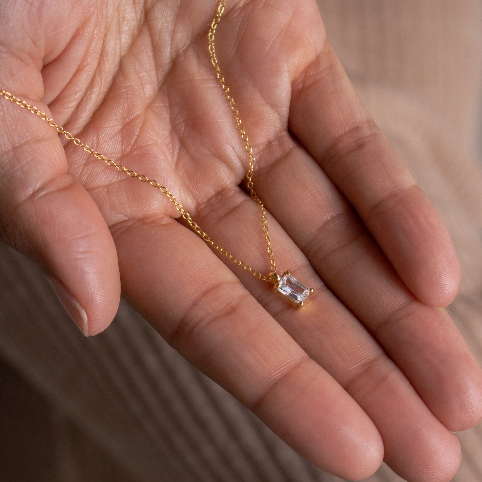 A hand holds the Sage Diamond Necklace, featuring a delicate gold chain with a rectangular diamond pendant, capturing the timeless elegance of luxury jewelry.