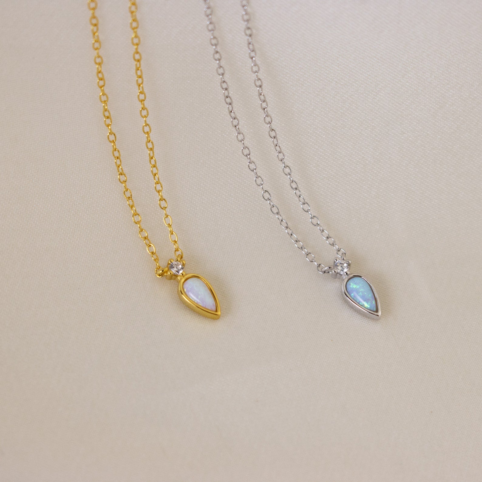 The Opal Drop Necklace, available in gold and silver, features an iridescent opal pendant. Shown on a beige fabric background, each necklace offers a timeless touch of elegance.