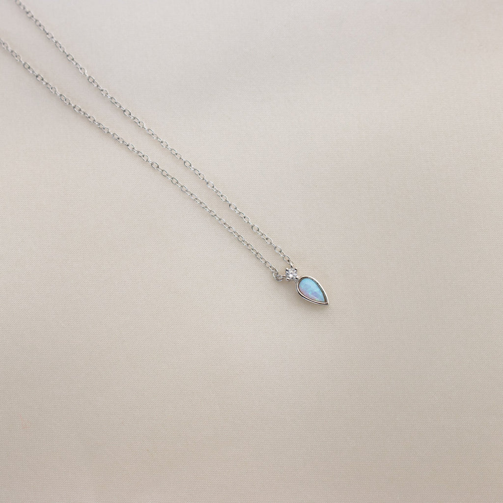 Opal Drop Necklace with a delicate silver chain and an iridescent opal teardrop pendant, shown on a light beige background.
