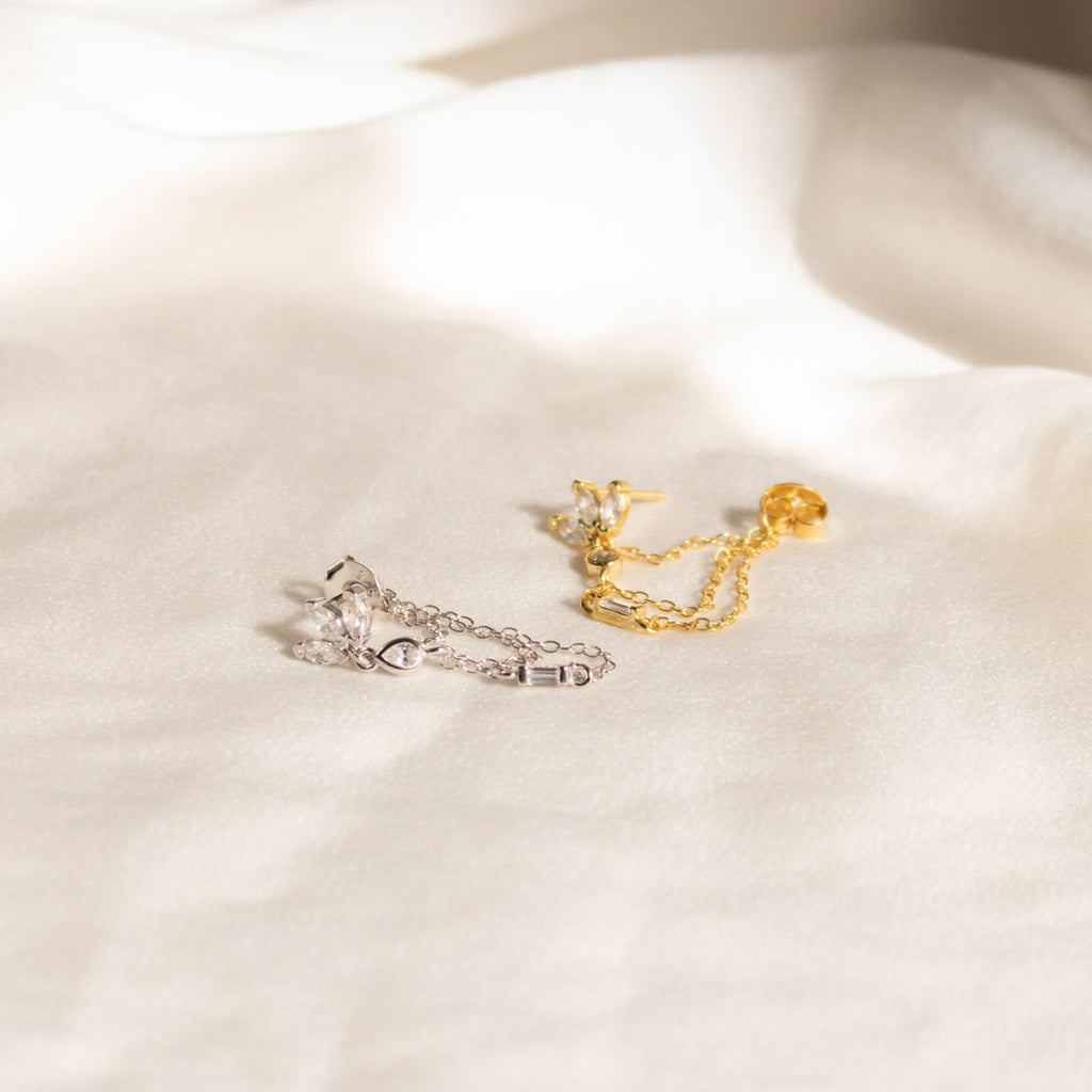 Genesis Chain Earrings in gold and silver, adorned with gemstones, make a stunning statement and are elegantly displayed on a soft white fabric background.