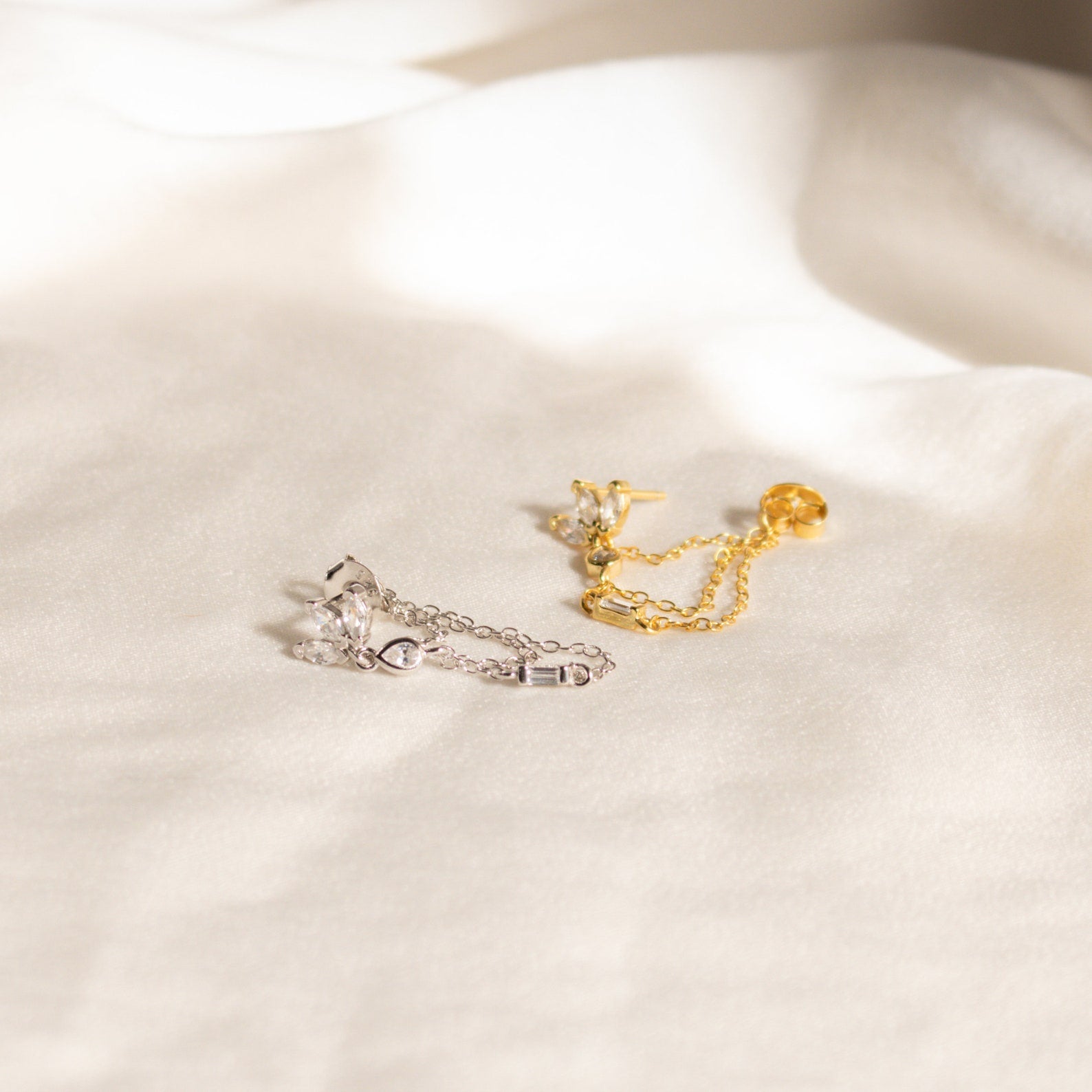 Genesis Chain Earrings in gold and silver, adorned with gemstones, make a stunning statement and are elegantly displayed on a soft white fabric background.