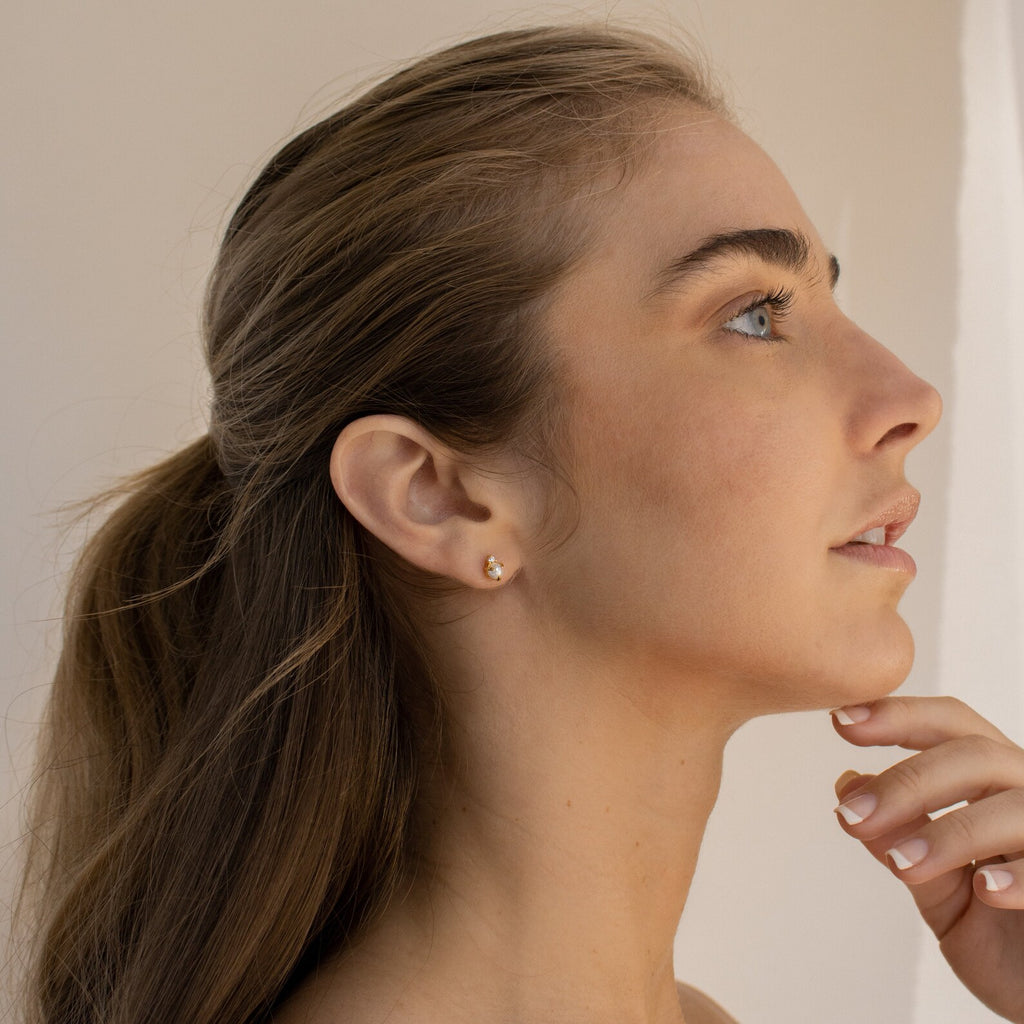 A woman with long brown hair in a ponytail, wearing Julia Pearl Studs, looks thoughtfully upward.