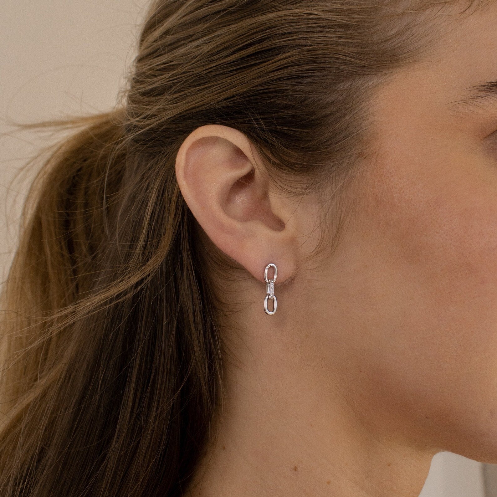 A woman with brown hair wears the Amia Pave Link Earrings, featuring a paperclip link chain design and pavé accents, shown in a side profile close-up.