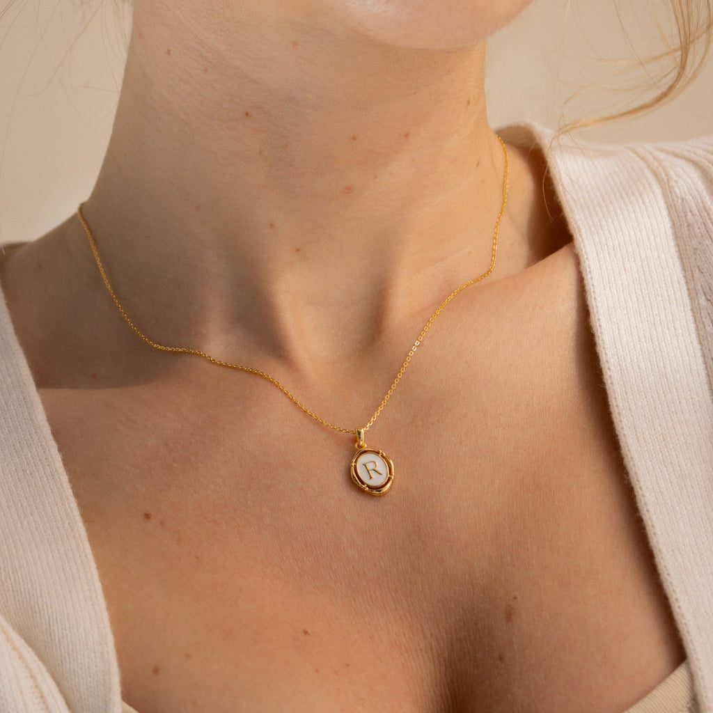 A woman in a cream top wears the Initial Pendant Necklace in 18K Gold—a vintage-inspired gold enamel necklace with a round pendant engraved with the letter R.