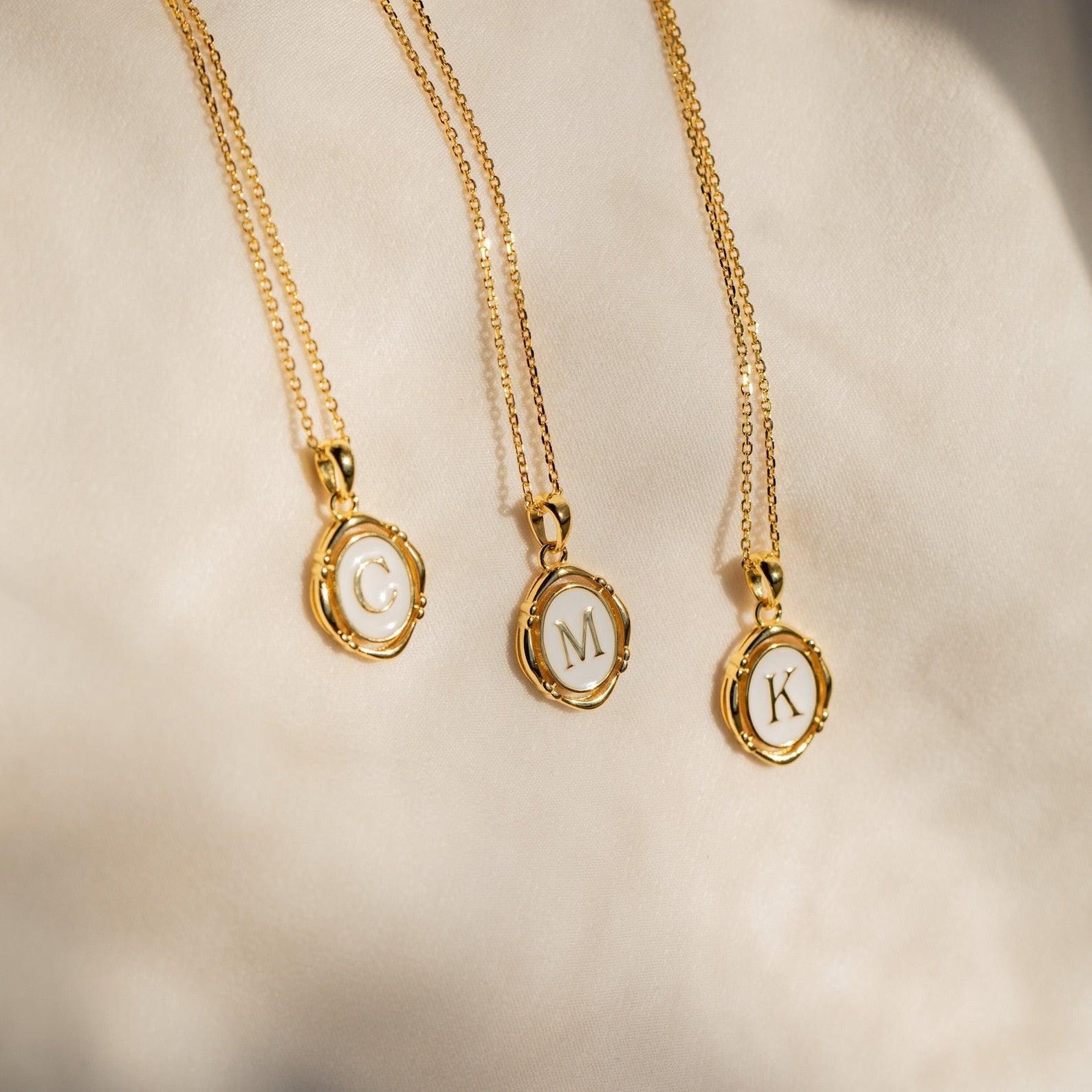 Three Initial Pendant Necklaces in 18K Gold, featuring round C, M, and K charms, are displayed on a soft beige fabric background, adding timeless vintage style to any outfit.