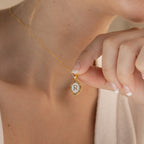 A gold chain necklace with a white oval pendant featuring the letter 'R' in gold lettering with the pendant being held to the camera by a model.