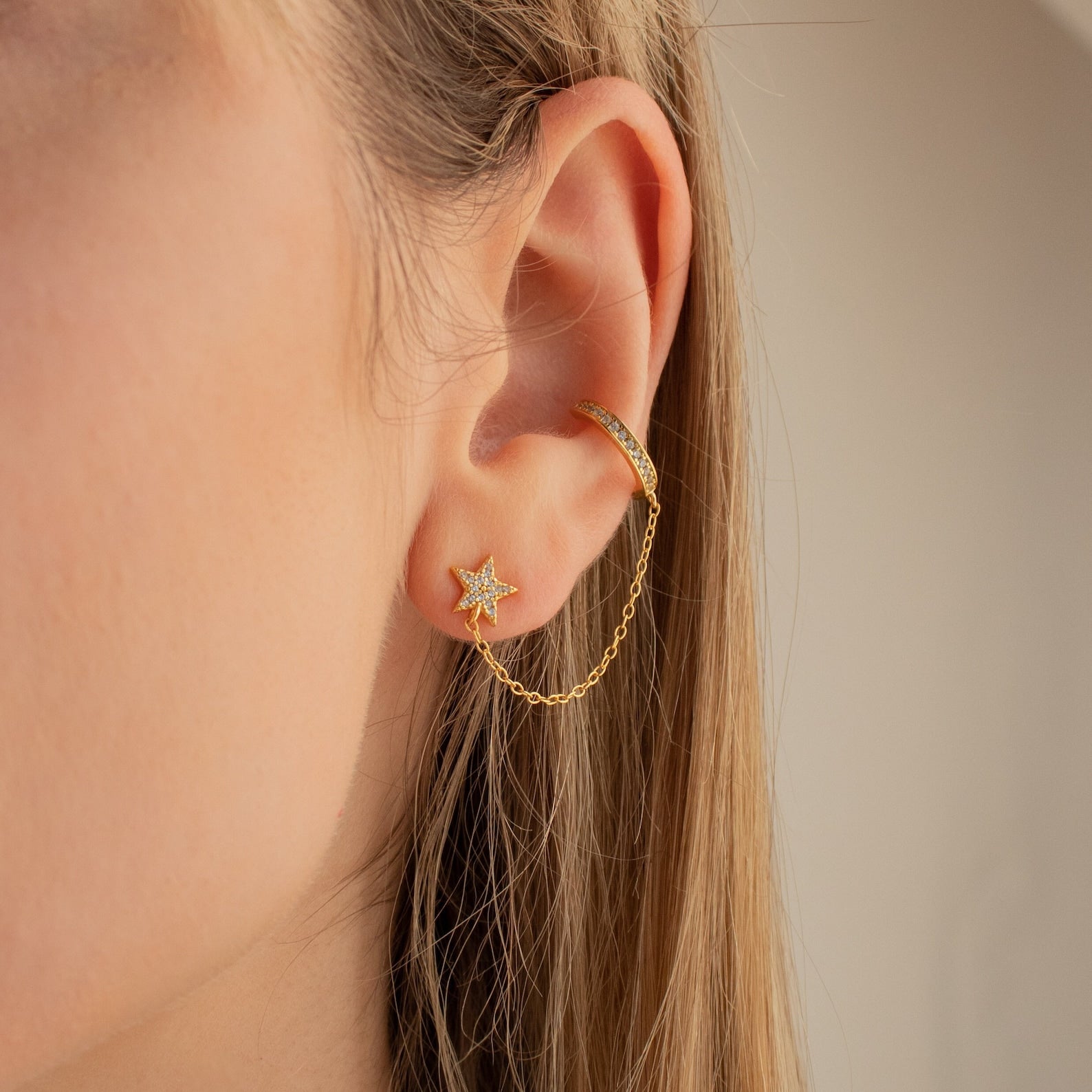 Close-up of Star Pave Chain Ear Cuffs, featuring a diamond-studded star charm linked by a chain to an adjustable cuff, worn on an ear with light brown hair.