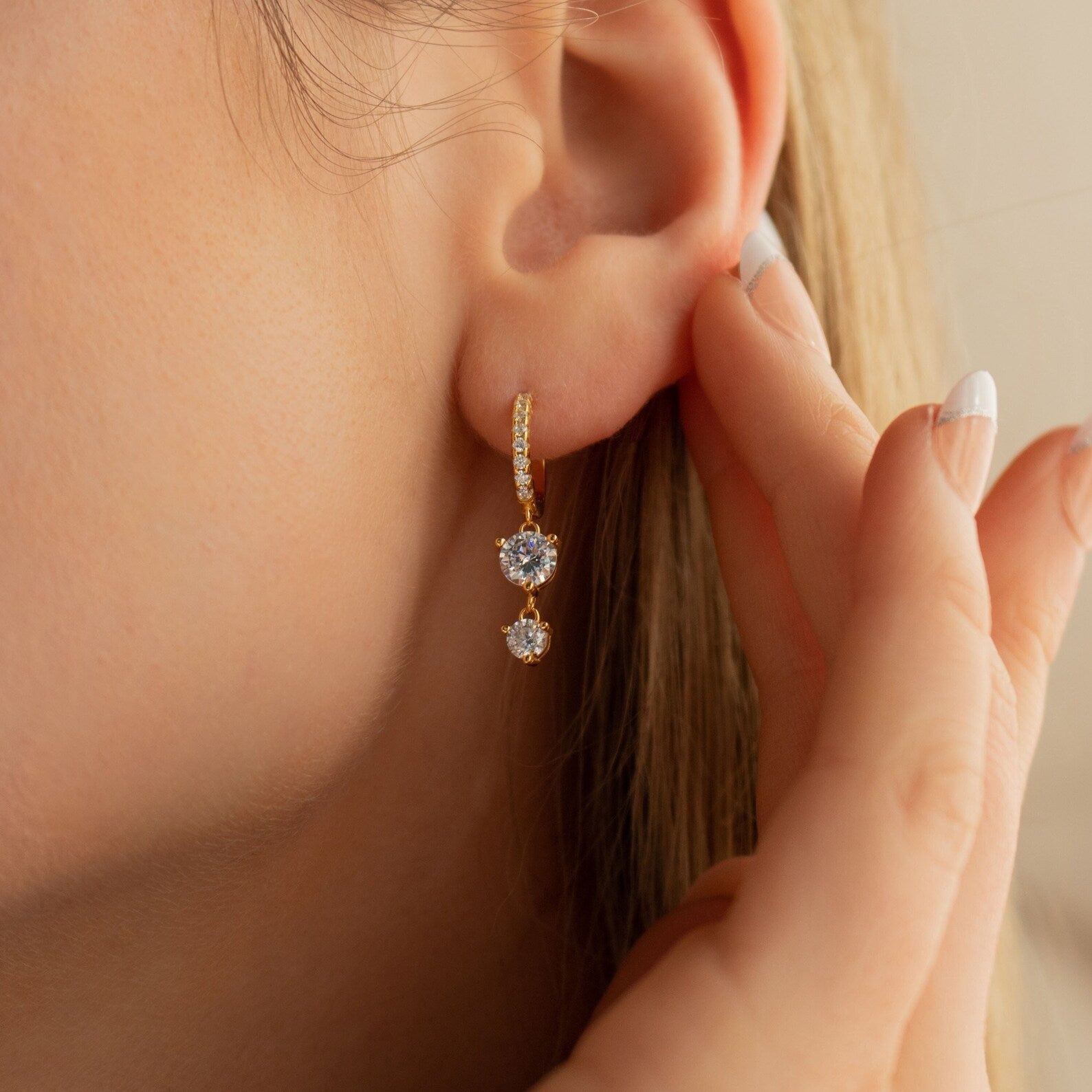 Close-up of a woman’s ear wearing Caroline Pave Drop Huggies featuring diamond hoops with two blue gemstones, as her hand gently touches her ear.