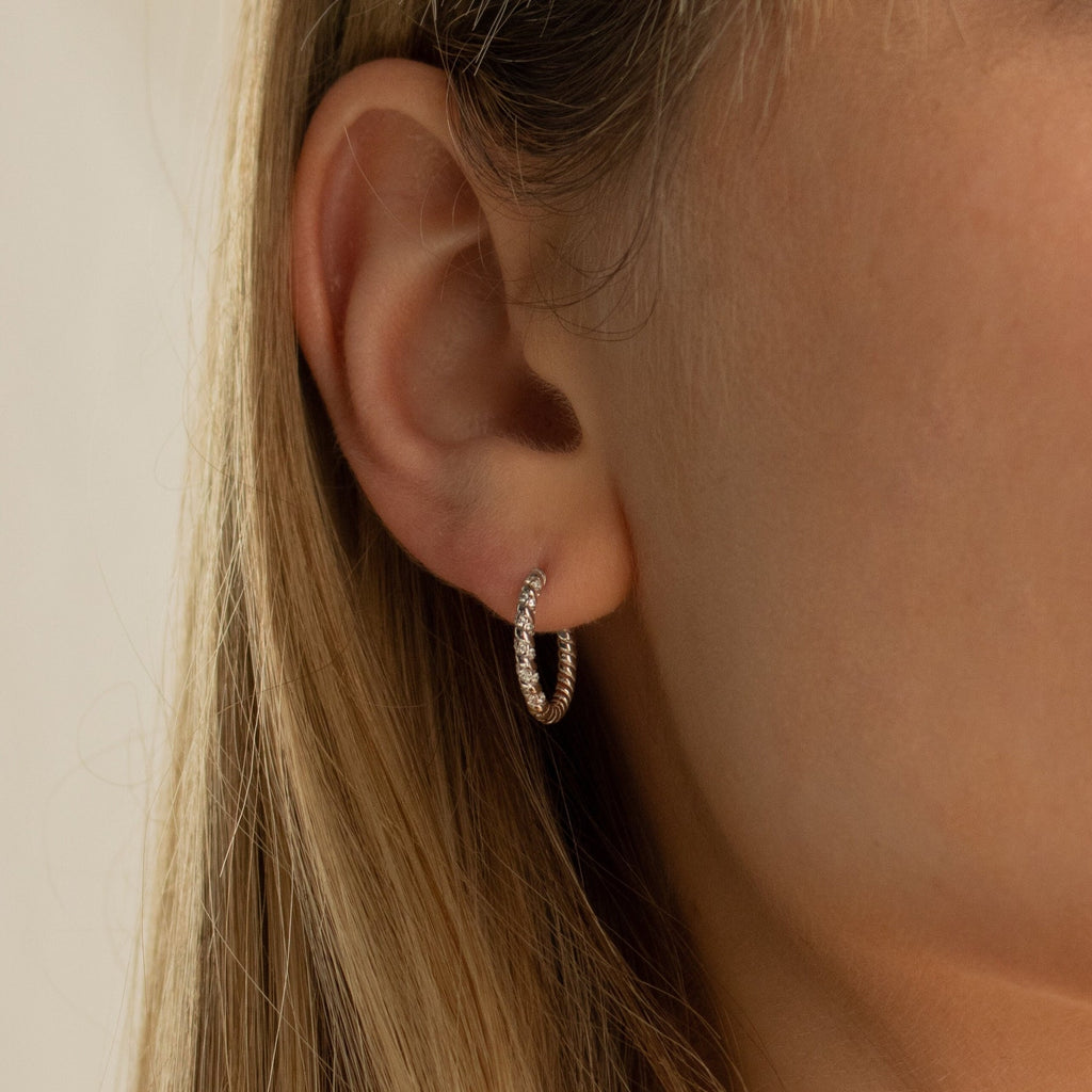 A close-up of a woman's ear adorned with Aster Pave Twisted Hoops—delicate gold hoops featuring embedded stones and an elegant twisted design.