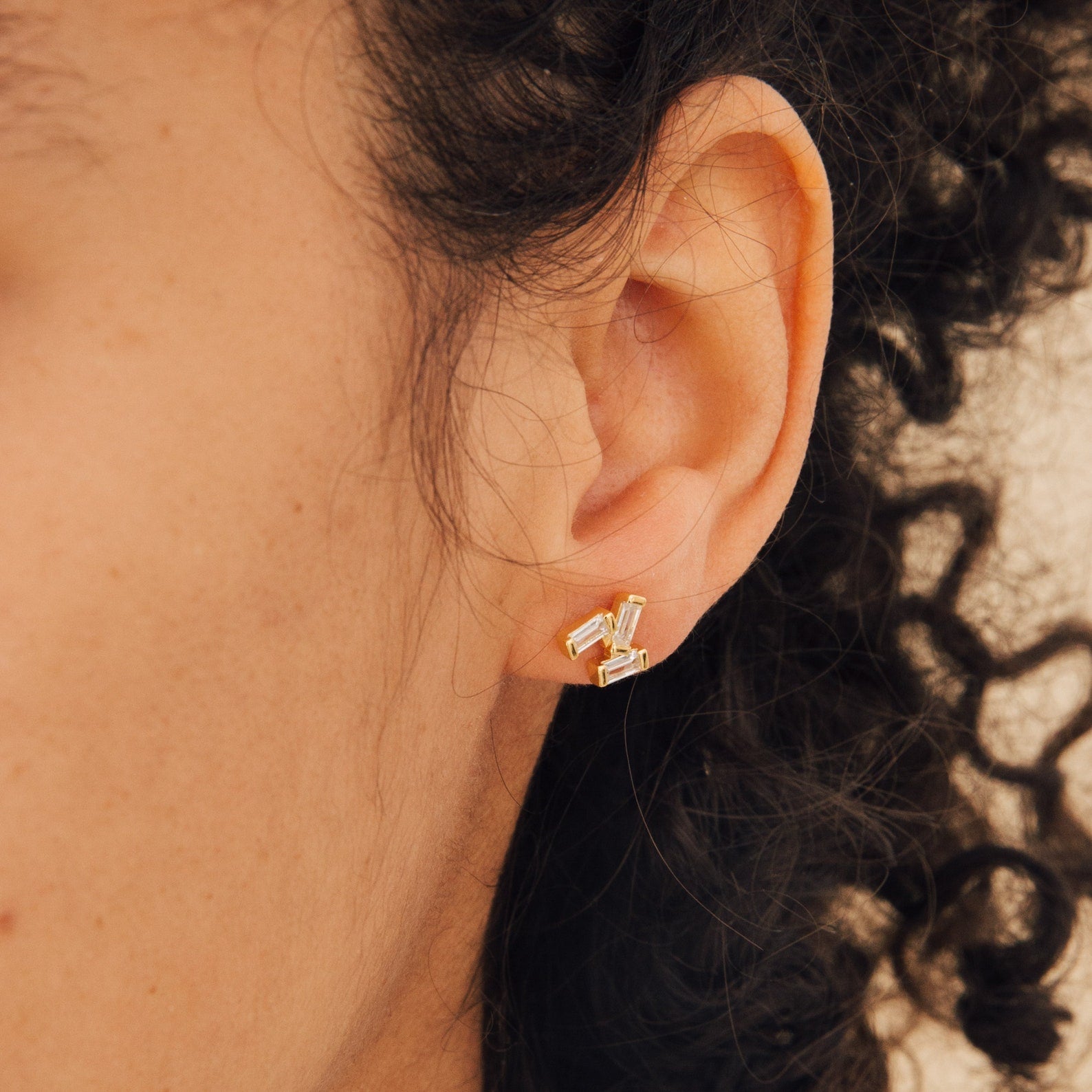 Close-up of a woman's ear wearing Glacier Diamond Studs, featuring small gold earrings set with rectangular gemstones.